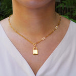 Gold chain link necklace with a lock pendant worn by a person in a white top, with a blurred green background.